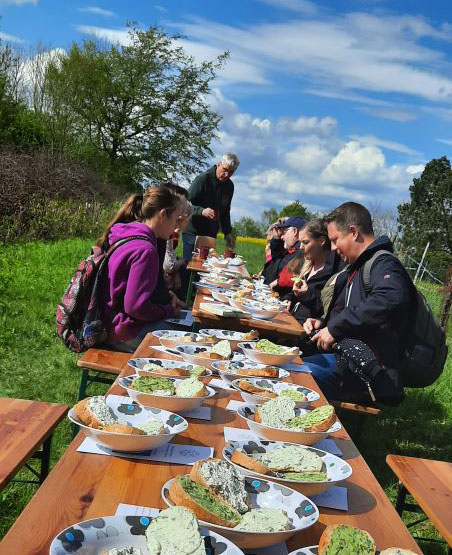  Teilnehmer der Kräuterwanderung genießen zum Abschluss selbstgemachte Kräuterdips und Kräuterbutter in der idyllischen Frühlingslandschaft.